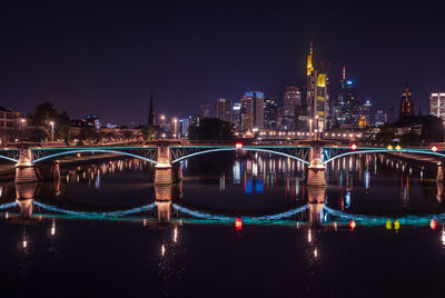 Illuminated bridge over river by buildings against sky at night