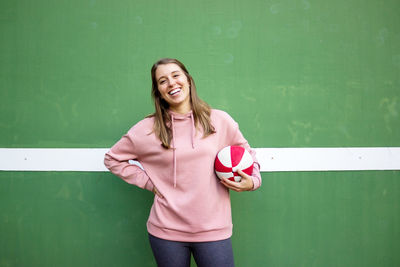 Portrait of smiling young woman holding basketball standing against wall