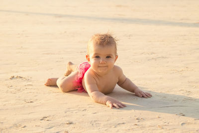 Little baby girl is lying on a sandy beach near to sea in sunset sunlight.