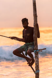 Full length of man sitting on shore against orange sky
