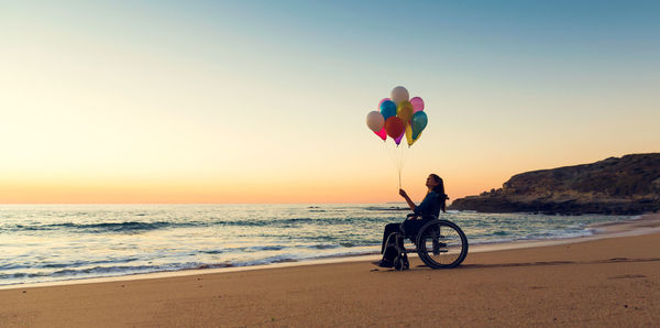 Man with balloons at beach against sky during sunset