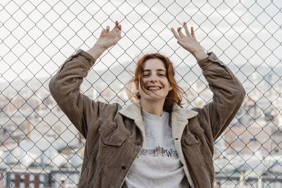 Young beautiful woman smiling looking at camera in front of metal fence with city view behind