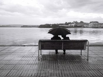 Rear view of couple sitting with umbrella on bench at promenade against cloudy sky