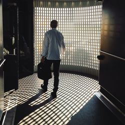 Rear view of man carrying bag while walking in building by window