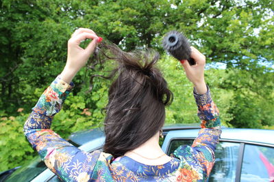 Rear view of woman combing hair by car against trees