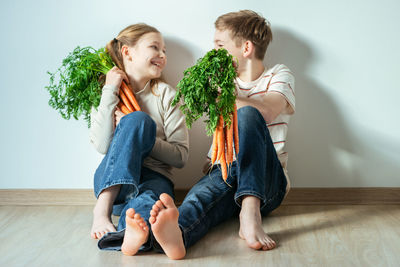 Smiling sibling holding carrot while sitting against wall