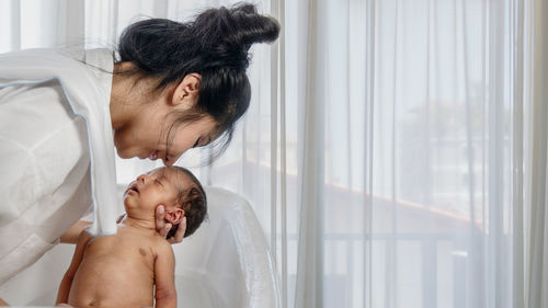 Side view of woman holding naked newborn son against curtain at home