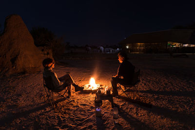 Man and woman sitting on chairs by campfire at night