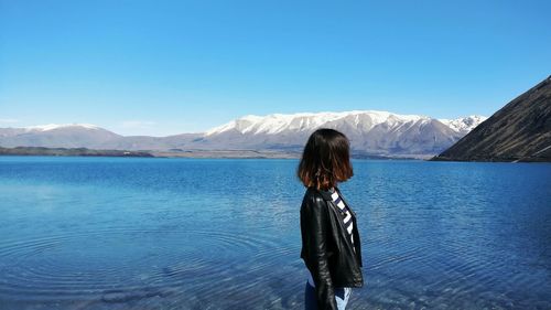 Side view of woman standing by lake against mountains and clear blue sky