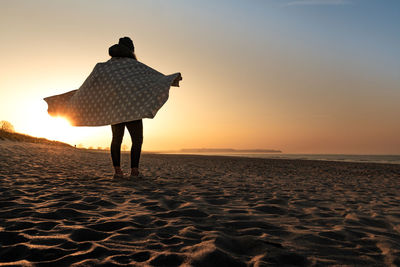 Rear view of woman standing at beach against sky during sunset