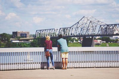 Rear view of woman and man standing by railing against cloudy sky