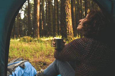 Side view of woman looking up while holding coffee mug in tent at forest