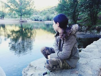 Young woman sitting by lake against trees