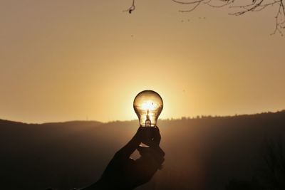 Cropped hand holding light bulb against sky during sunset