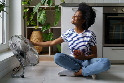 Heat-tired african american woman catching cold air of fan sits on floor with happy expression