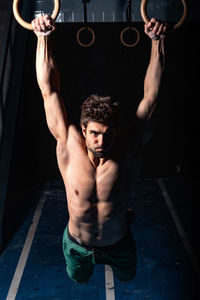 Full length strong shirtless guy looking at camera doing exercise on gymnastic rings during intense workout in dark gym