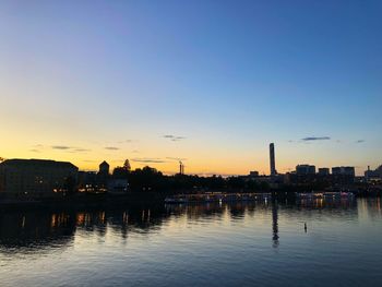 Silhouette buildings by river against clear sky at sunset