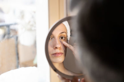 Crop young woman in white headband applying eyeshadow with hand and looking at reflection in mirror while sitting in front of large window