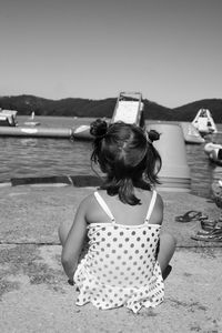 Rear view of girl sitting by lake against clear sky