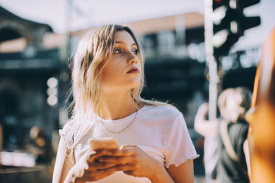 Young woman looking away while using mobile phone on street in city