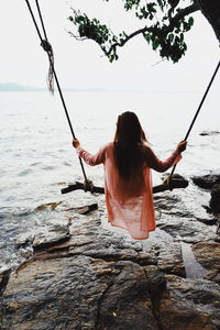 Rear view of woman sitting on swing against sea and sky