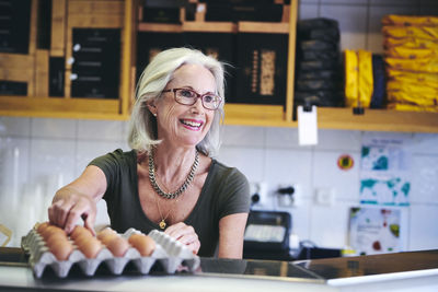 Senior saleswoman smiling while picking brown egg from carton in deli