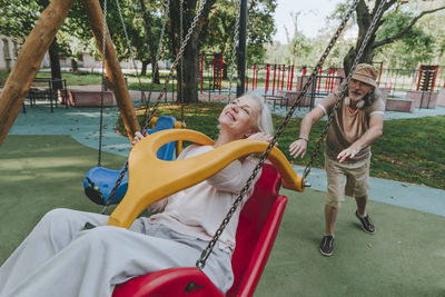 Senior man pushing woman enjoying swing at park