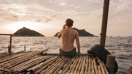 Rear view of shirtless man sitting on pier over sea against sky during sunset