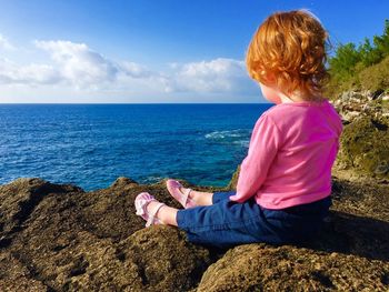 Full length of girl sitting on rock by sea against sky