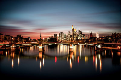 Illuminated bridge over river by buildings against sky in city