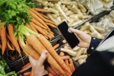 Cropped image of woman using phone while buying carrots in supermarket