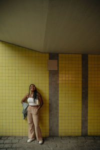 Full length of smiling teenage girl standing with hands in pockets against yellow wall