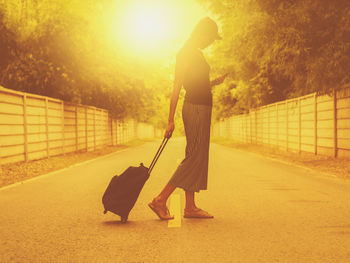 Side view of woman pulling suitcase on road amidst trees during sunset