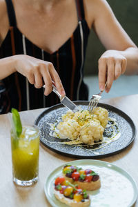 Crop woman cutting cauliflower in sauce at table with sandwiches and fresh lemonade in restaurant