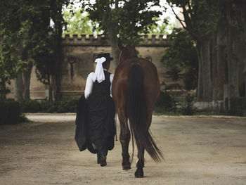 Full body of african american adult lady in elegant clothes and hat walking with brown horse near trees and building in daytime