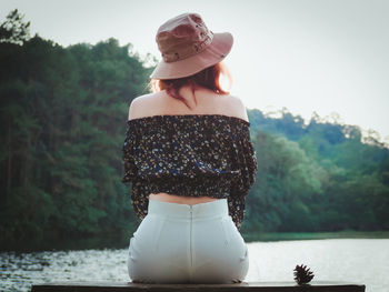 Woman wearing hat standing by lake against trees