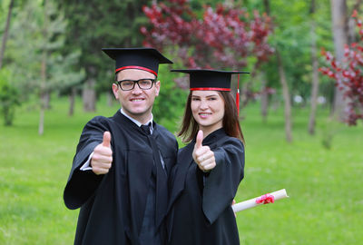 Portrait of couple wearing graduation gown showing thumbs up sign on field