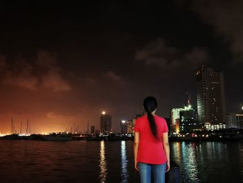 Rear view of woman standing by lake against illuminated city