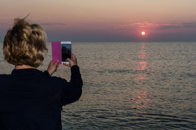 Rear view of woman photographing sea against sky during sunset