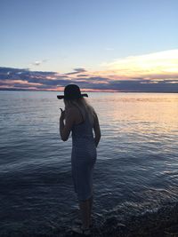 Rear view of teenage girl standing by sea against sky during sunset