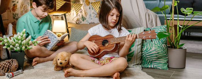 Banner of little girl playing ukulele while boy reading book on handmade teepee in living room