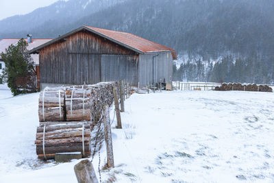Firewood on a barn in snowy bavarian landscape in snowfall and mountain forest in the background.