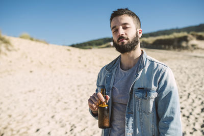 Young man holding alcoholic drink bottle while standing at beach during sunny day