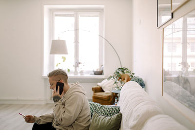 Side view of woman with pregnancy test results talking through smart phone while sitting on sofa in living room at home