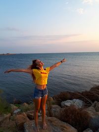 Woman with arms outstretched standing on rocky shore against sky