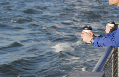 Cropped hands of man holding disposable cup while leaning on railing by sea