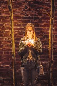 Portrait of young woman holding illuminated string light against brick wall at night