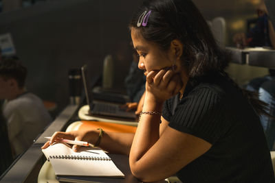 Young female student with hand on chin reading book while sitting in lecture hall at university