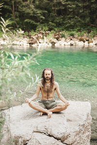 Shirtless young hipster meditating in lotus position on rock