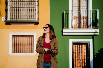 Smiling woman with map standing against wall during sunny day